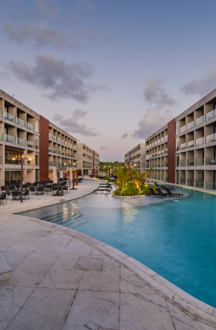 A imagem mostra uma vista de um hotel moderno, com uma piscina ao centro e rodeada por edifícios de vários andares. A água da piscina reflete a luz suave do final da tarde, criando uma atmosfera tranquila e relaxante. Espreguiçadeiras estão dispostas ao redor da piscina, e pequenas plantas decoram a área. O céu, em tons de laranja e azul, indica que a imagem foi tirada durante o entardecer. O cenário transmite uma sensação de conforto e sofisticação.