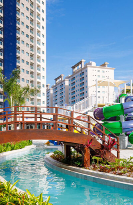 A imagem mostra uma piscina com um pequeno canal, atravessado por uma ponte de madeira. Ao fundo, há um conjunto de edifícios modernos com varandas e várias janelas. À direita, um grande conjunto de toboáguas coloridos em tons de verde, roxo e azul se destaca, sugerindo uma área de lazer aquático. O céu está claro e ensolarado, criando um ambiente alegre e descontraído.