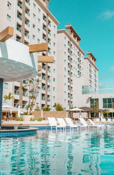 A imagem mostra uma piscina em primeiro plano, com várias espreguiçadeiras brancas ao redor. No fundo, há um grande edifício moderno com várias janelas. O céu é claro e azul, e a luz suave sugere um dia de sol. A cena transmite um ambiente de descanso e lazer, típicos de um resort ou hotel.