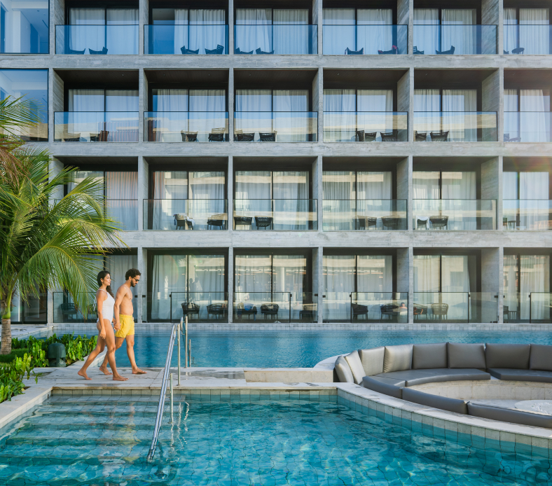 Casal caminhando ao lado de uma ampla piscina em um resort de luxo com varandas modernas, rodeado por palmeiras e ambiente tropical, ideal para férias de lazer e relaxamento.