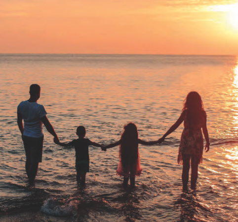 Família aproveitando o pôr do sol à beira-mar. O casal caminha de mãos dadas com duas crianças dentro da água, enquanto o sol se põe no horizonte, tingindo o céu e o mar com tons dourados e alaranjados. As ondas suaves tocam seus pés, criando uma atmosfera de paz e conexão. A cena transmite um momento de união, amor e tranquilidade em um cenário paradisíaco.