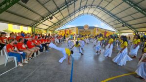 Apresentação cultural com capoeira e dança em evento dos Amigos do Bem, com voluntários assistindo em uma quadra coberta.