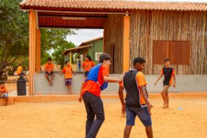 Jovens jogando futebol em um ambiente rústico ao ar livre, com construção de madeira ao fundo.