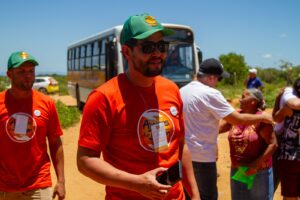 Voluntário dos Amigos do Bem com boné verde e camiseta laranja, segurando um celular, enquanto interage em uma ação social ao ar livre.